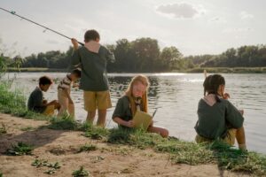 Kids engaged in fishing and reading by a serene lakeside during daylight, wearing scout uniforms.